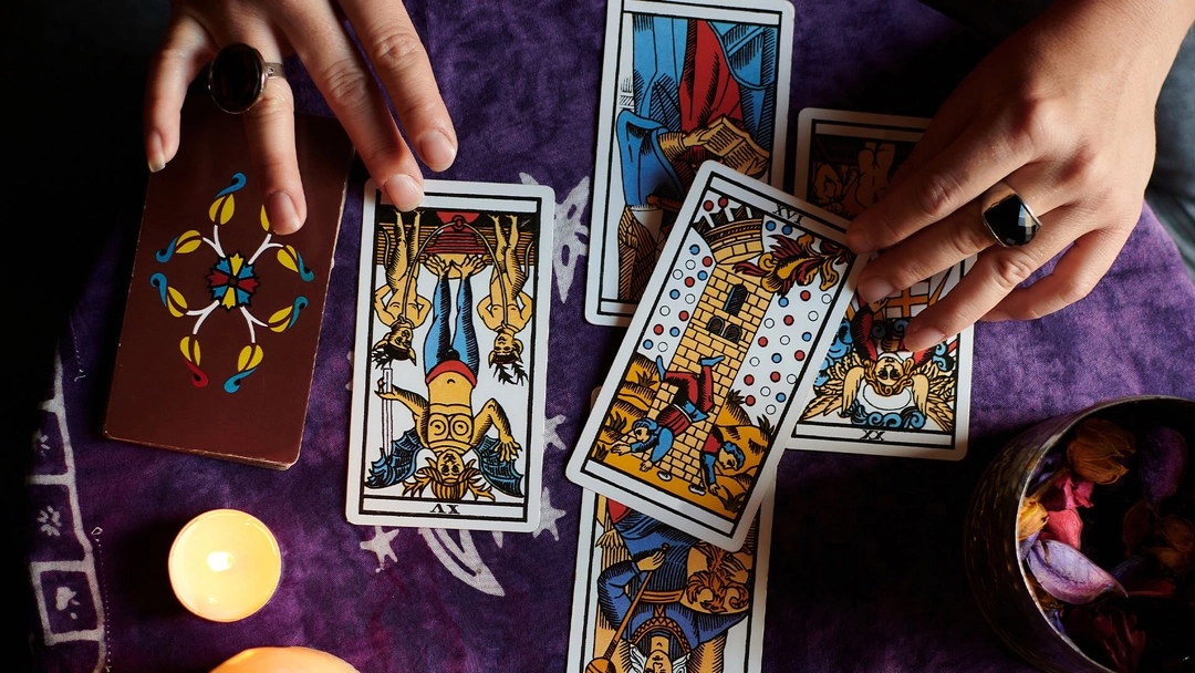 Hands with rings displaying several tarot cards face up on a purple cloth next to a lit candle and a bowl with dried petals.